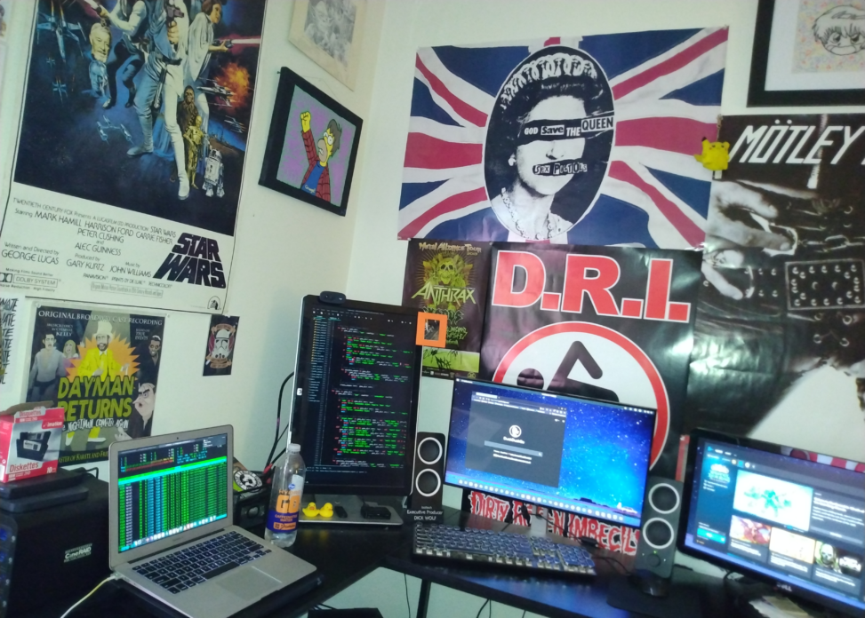 A desk with three monitors and a laptop. Posters are on the wall behind it.