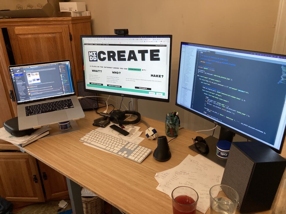 A laptop and two screens on a standing desk. Papers, cords, two drinks, some mints are on the desk.